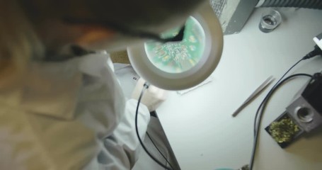 Young energetic female tech using lamp with magnifying glass to repair electronic equipment in research facility - Powered by Adobe