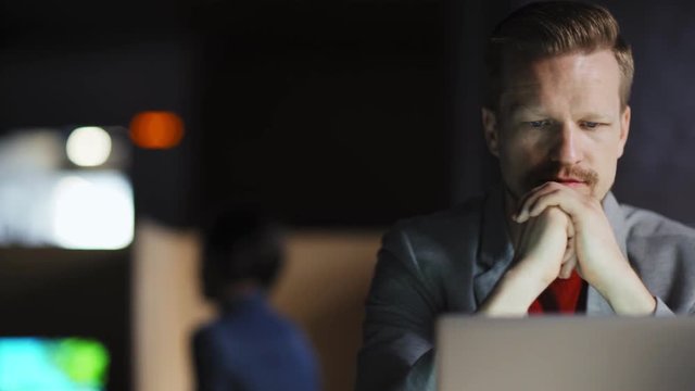 Tracking shot of middle aged businessman sitting at office desk at night, reading document on laptop screen with his hand on chin and thinking
