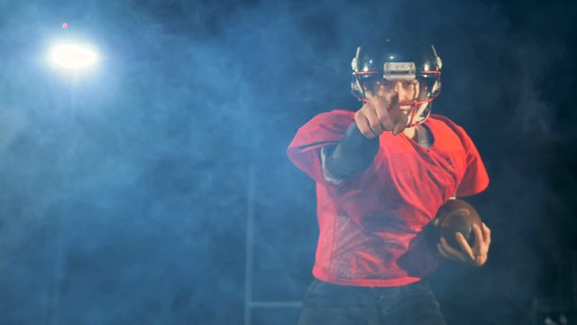 American football athlete is running with a ball and pointing into the camera
