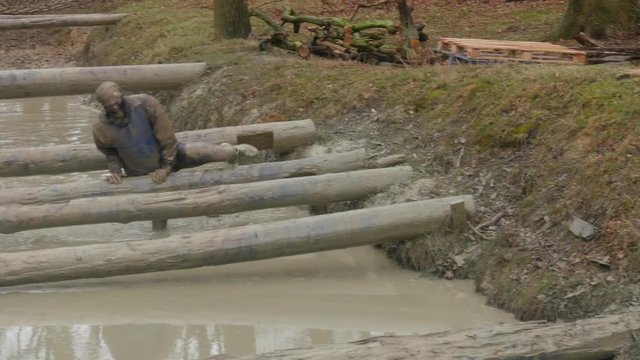 An obstacle course racer climbs over logs while in a muddy stream