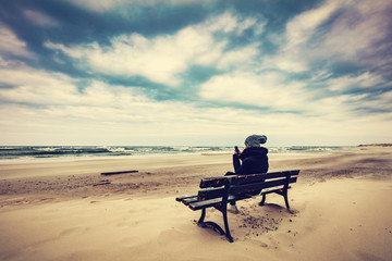 Woman sitting on a bench on the beach using her phone - obrazy, fototapety, plakaty