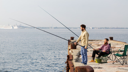 leisure and people concept - male friends with fishing rods on pier at sea