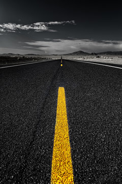 A black and white image with a yellow line in the centre in the middle of a road heading straight into the endless distance.