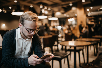european male businessman with glasses uses a smartphone in a coworking cafe with a warm cozy light