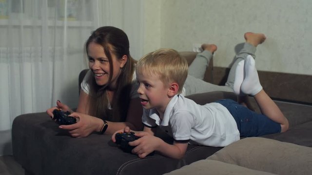 Emotional portrait of mom and son at home on the couch, they play an interesting video game with a joystick in their hands. High resolution.