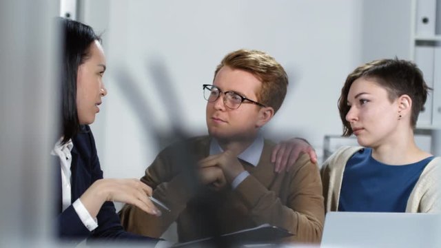 Hidden camera handheld shot with bokeh of 30-something Caucasian husband and wife getting professional legal advice from Asian female lawyer in her office, attentively listening to her and nodding