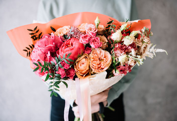 very nice young woman holding a beautiful blossoming flower bouquet of fresh peonies, roses, eustoma, pistachio, carnations in tender pink and vivd magenta colors on the grey wall background