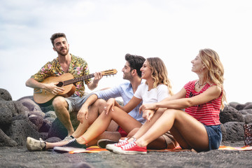 Group of young friends having good time at the beach playing guitar and singing songs at sunset. Youth lifestyle concept