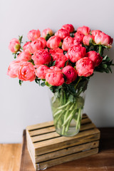 Big blossoming beautiful coral peonies mono bouquet in a glass vase standing on a wooden rustic box on the grey wall background 