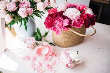 Obraz na płótnie Canvas Beautiful blossoming fresh peonies in a wicker basket , tender pink peony bouquet in a vase and coral peony petals on the pink table, summer floral setup