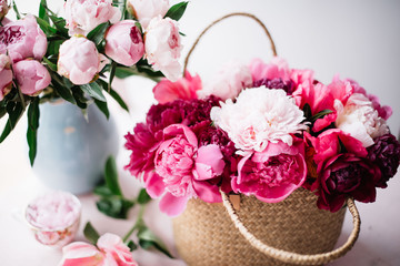 Beautiful blossoming fresh peonies in a wicker basket , tender pink peony bouquet in a vase and coral peony petals on the pink table, summer floral setup