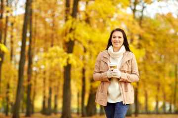 season, hot drinks and people concept - happy young woman drinking takeaway coffee or tea from disposable paper cup at autumn park