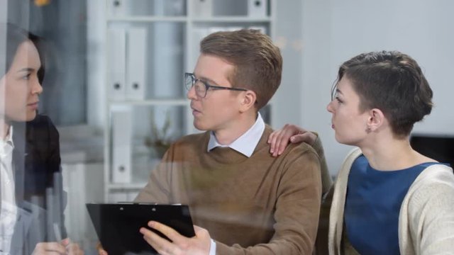 Hidden camera tracking shot of young Caucasian man and woman sitting in solicitors office, holding clipboard with document draft and discussing it with professionally dressed Asian female legal expert