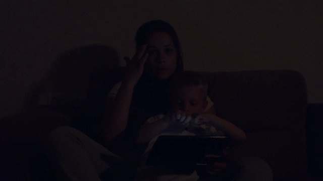 Cute little boy playing tablet with mom at night in bed in the dark. Portrait of a mother with her son with a tablet at home in the dark, the light from the screen shines on their faces. 4K.