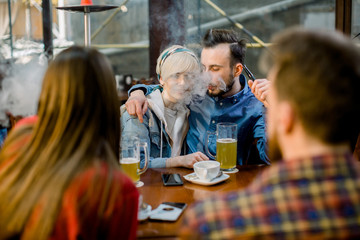 People Meeting Communication Concept. Group of multiethnical students sitting in a cafe bar having fun. Young cheerful couple hugging, kissing and smoking hookah