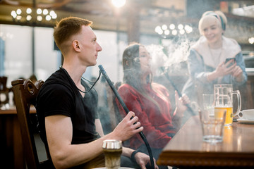 Caucasian young man in black t-shirt smoking a hookah while sitting at the table in cafe indoors together with friends
