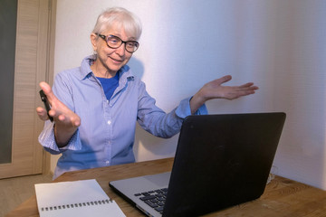 An elderly caucasian woman wearing glasses, wearing a blue-and-white striped shirt and a blue T-shirt, happily exclaims and looks at the monitor screen. She rejoices in winning or success.