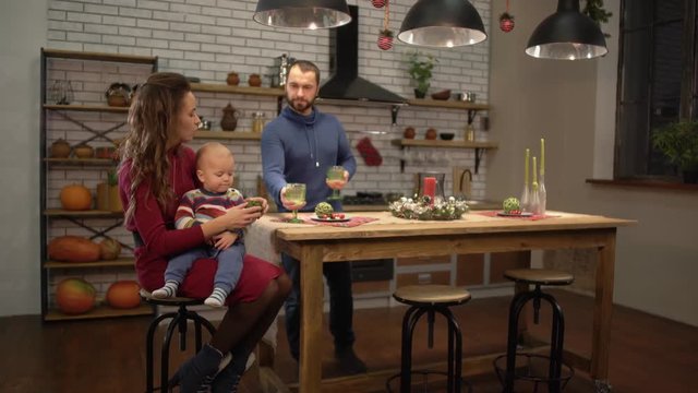 Young pretty woman sitting at the table in the kitchen with baby on her pals while handsome bearded man bringing her glass of lemonade drink. Happy family spend time together