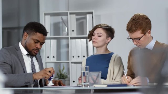 Waist-up shot of young Caucasian man and woman each signing separate copies of document, then African American male notary sealing them with self-inking rubber stamp, and continuing conversation