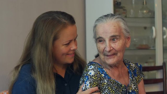 A young woman hugs her ninety year old grandmother at home on the couch. Granddaughter came to visit an old grandmother. An old woman with gray hair and wrinkles talking to an adult granddaughter.