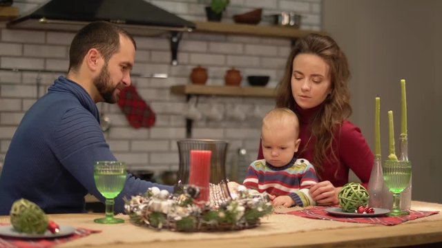 Mother, father and a baby have fun in modern kitchen. Dad and mom playing with child, man spinning round candy on the table to entertain a boy. Happy family spend time together