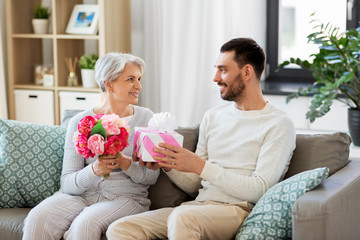 family, mother's day and birthday concept - smiling adult son giving present and flowers to his...