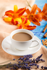 Orange day-lily and lavender flowers and a cup of coffee on a blue concrete background, with orange textile. side view.