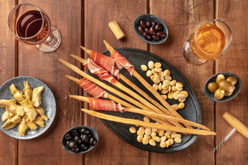 Italian antipasti. Grissini breadsticks with parma ham and roasted almonds, with olives and artichokes, shot from the top on a dark rustic wooden background with wine glasses