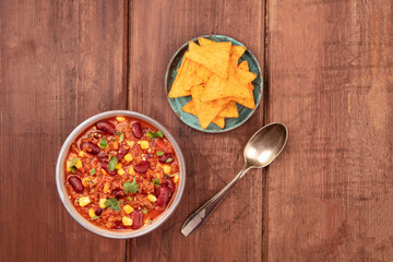 Chili con carne, a traditional Mexican dish with red beans, cilantro leaves, ground meat, and chili peppers, shot from the top with nachos on a dark rustic wooden background