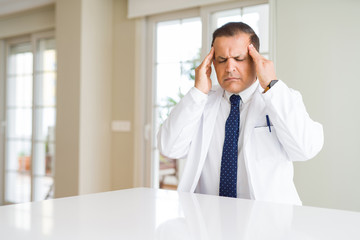 Middle age doctor man wearing medical coat at the clinic with hand on head for pain in head because stress. Suffering migraine.