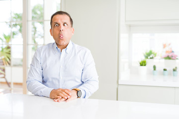 Middle age man sitting at home making fish face with lips, crazy and comical gesture. Funny expression.