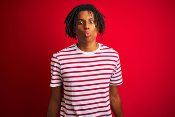 Young afro man with dreadlocks wearing striped t-shirt standing over isolated red background making fish face with lips, crazy and comical gesture. Funny expression.