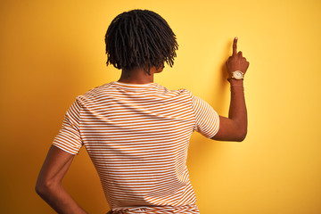 Afro man with dreadlocks wearing striped t-shirt standing over isolated yellow background Posing backwards pointing ahead with finger hand