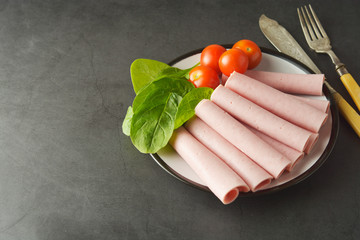 Thin slices of ham rolled on plate with fresh vegetables, dark background. Breakfast food,...