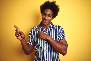 American man with afro hair wearing striped shirt standing over isolated yellow background smiling and looking at the camera pointing with two hands and fingers to the side.