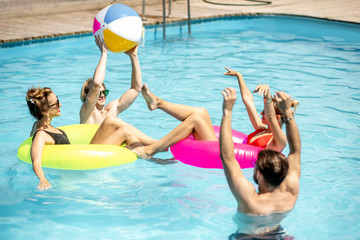 Group of a happy friends having fun, swimming with inflatable rings and playing with ball in the swimming pool outdoors during the summertime