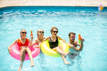 Portrait of a happy friends standing in the swimming pool with inlatable rings and water guns, enjoying summertime outdoors