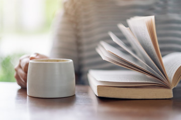 Closeup image of a woman holding and reading a book while drinking coffee on wooden table