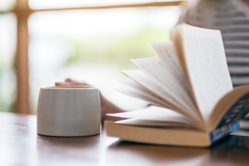 Closeup image of a woman holding and reading a book while drinking coffee on wooden table