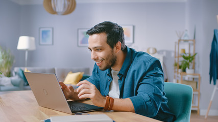 Portrait of Handsome Young Man Using Laptop Computer at Home, Watching and Smiling at a Content. Cozy Modern Flat done in Scandinavian Style.