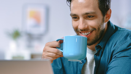 Portrait of Focused Young Man Working on a Laptop from Home, He Drinks Beverage From the Mug and is Satisfied with His Work. Cozy Modern Flat done in Scandinavian Style.