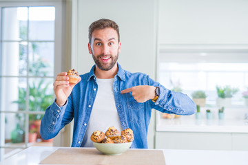 Handsome man eating chocolate chips muffin with surprise face pointing finger to himself