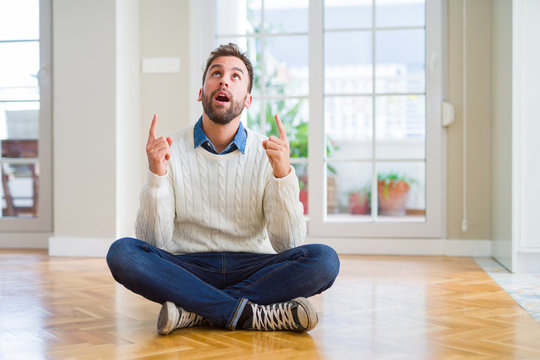 Handsome man wearing casual sweater sitting on the floor at home amazed and surprised looking up and pointing with fingers and raised arms.