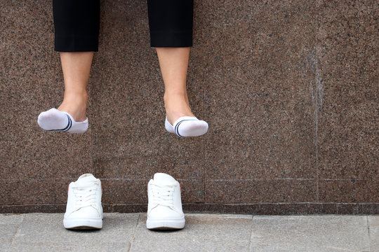 Tired legs, foot pain, barefoot woman with removed white sneakers resting on a street. Female legs in white socks dangling over shoes