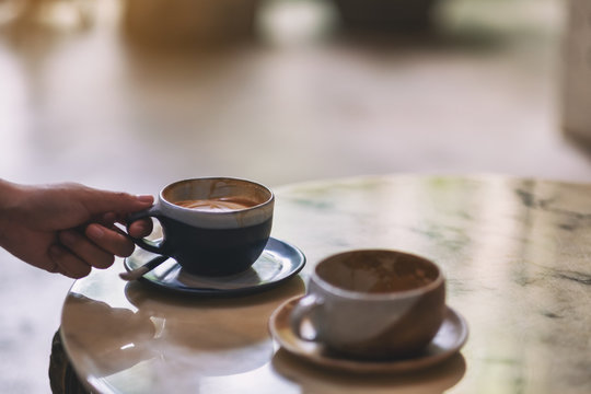 Closeup image of a hand holding a blue cup of hot coffee with another cup on marble table in cafe