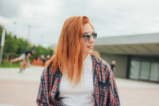 Attractive redhead smiling girl in round sunglasses in casual clothes sitting on street in city