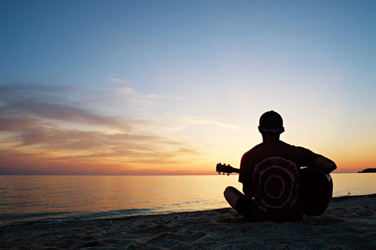 Young man wearing purple tie dye t-shirt playing dreadnought parlor acoustic guitar on beach at beautiful sunset time. Fit guitarist w/ sunburst instrument by the sea. Background, copy space, close up