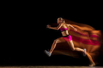 Professional female relay racer training isolated on black studio background in mixed light. Woman in sportsuit practicing in running. Healthy lifestyle, sport, workout, motion, action concept.