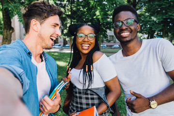 Education, lifestyle, leisure and technology concept. Cheerful african woman student in eyewear with two multiethnic men friends making selfie together in park during relax free time.