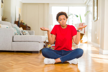 Young beautiful african american woman sitting on the floor at home clueless and confused...